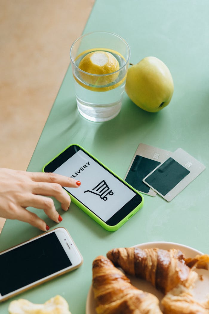 Hand using mobile app for food delivery. Croissant, apple, and credit cards on table.